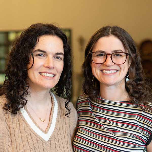 Two adults smile standing side by side indoors, facing the camera; one wears a beige sweater with a white V‑neck trim, and the other wears glasses and a striped top.
